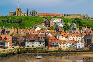 The red-roofed cottages of Whitby from across the harbour, with Whitby Abbey and Whitby parish church on the hill beyond